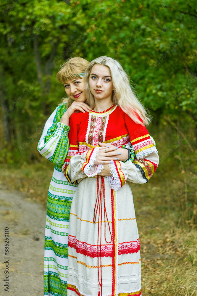 Two young women of different ages in folk costumes of red and green, are hugging each other in the park.