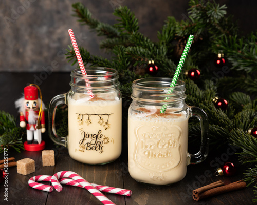 Traditional Christmas drink eggnog into glass mug from cocktail shaker. Cinnamon, anise stars on dark wooden table. Candy cane and fir-tree decoration photo