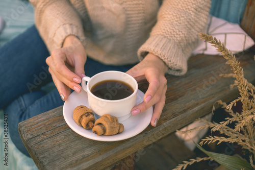 Autumn mood. A smiling girl in a cozy brown sweater and blue jeans sits on a bench and drinks coffee with croissants