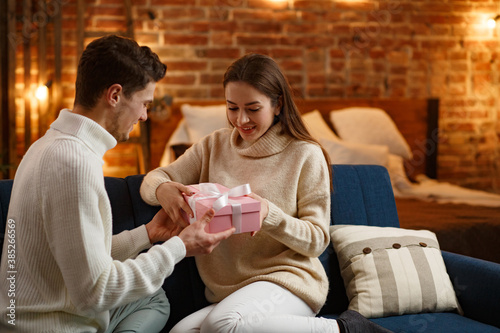 Handsome man presenting a gift to his beautiful girl and smiling. Beautiful young couple at home enjoying spending time together. Winter holidays, Christmas celebrations, New Year concept.