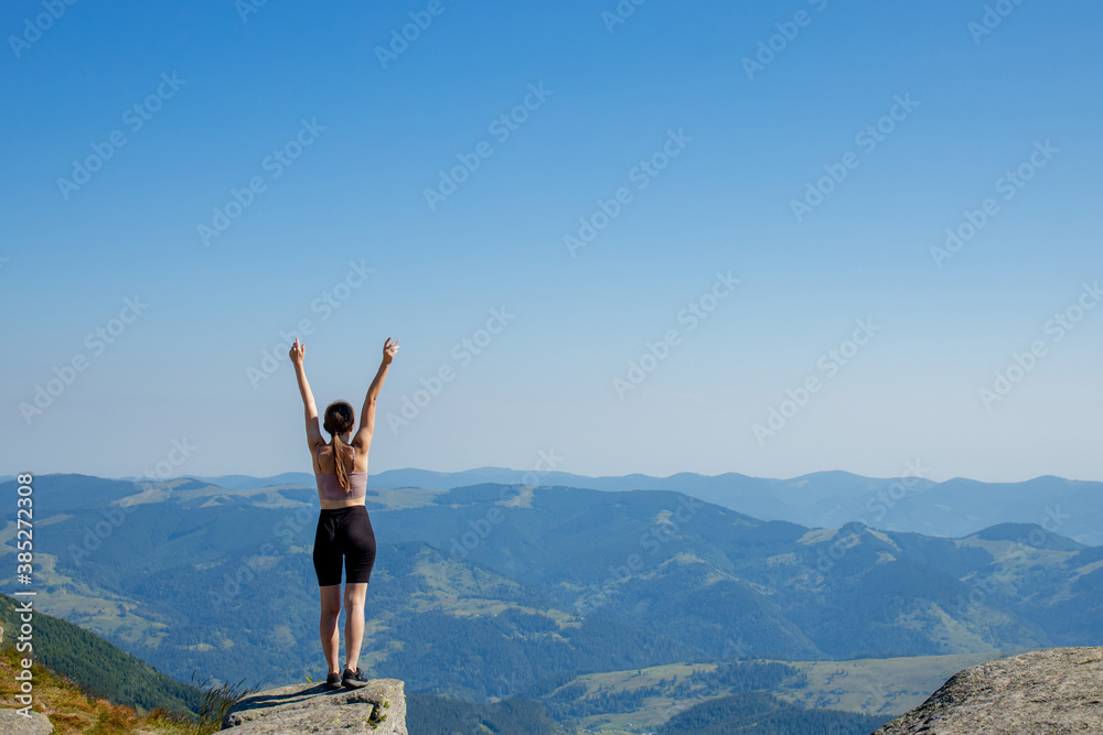 The young woman at the top of the mountain raised her hands up on blue sky background. The woman climbed to the top and enjoyed her success. Back view