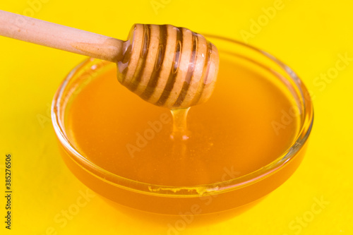  Honey in a glass bowl flows down the honey stick on a yellow background. Close-up.