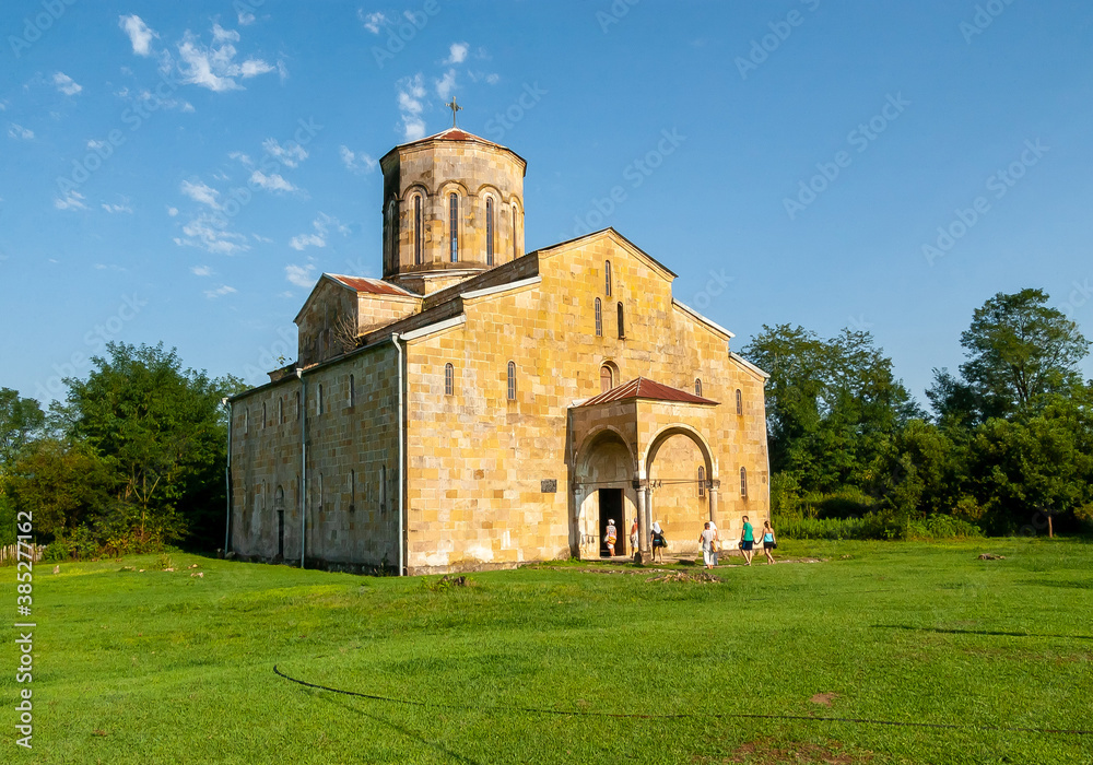 An ancient cross-domed cathedral made of yellow stone. Blue sky, green lawn, lush grass. At the entrance there are pilgrims and tourists. Abkhazia Mokva, Assumption Church 7th century.
