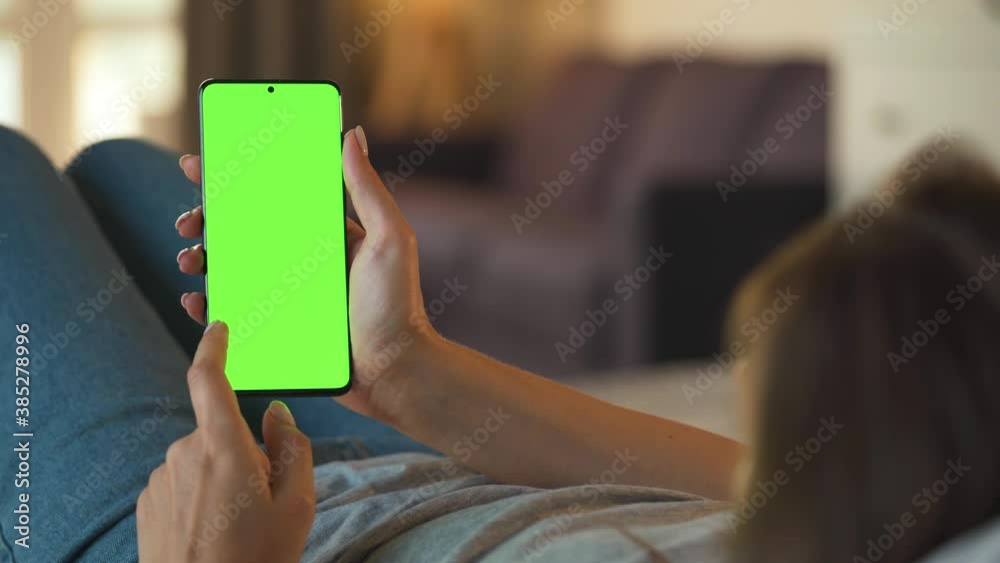 Woman at home lying on a sofa and using smartphone with green mock-up screen in vertical mode. Girl browsing Internet, watching content, videos, blogs. Woman at home lying on a sofa and using smartphone with green mock-up screen in vertical mode. Girl browsing Internet, watching content, videos, blogs.