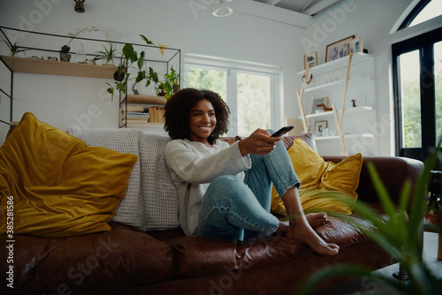 Beautiful young woman watching television sitting surrounded by cushions on a comfortable sofa with the remote control in her hand