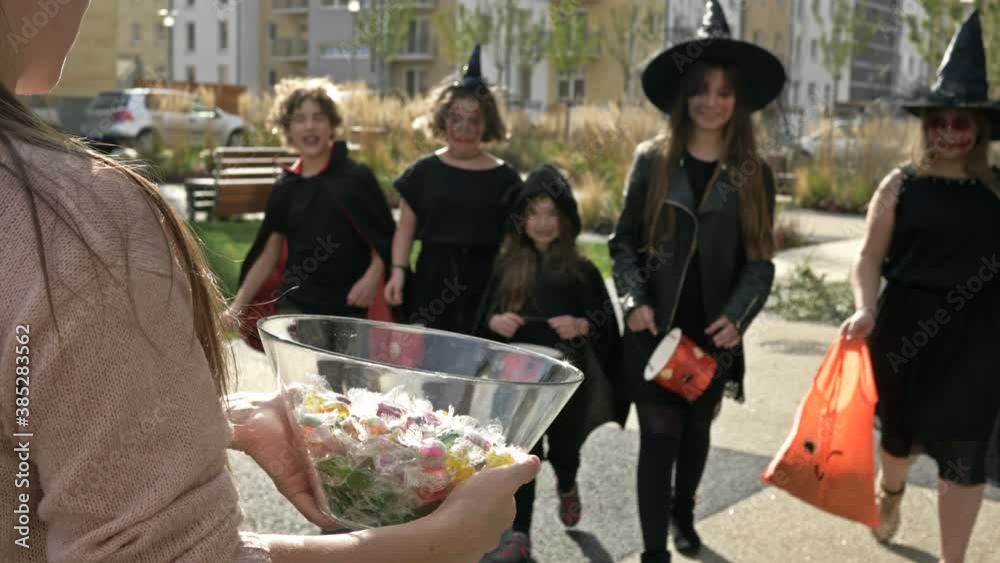 Woman treats sweets to a group of children in black fancy dress with scary makeup on their faces. Halloween traditions.