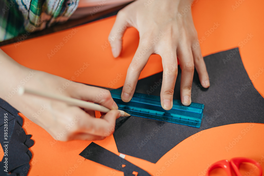 Overhead closeup shot of a woman drawing a line with a ruler on black paper. DIY Halloween arts and crafts as home activity for kids.