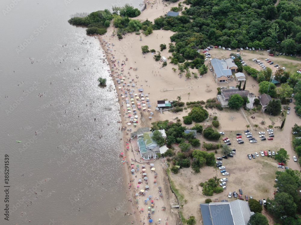 Foto de un dron, donde vemos la costa de un río con playas de arena, gente en el agua y personas con sombrillas en la costa.