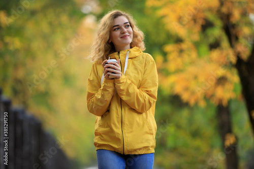 Woman in yellow jacket drinks a tea walking along fall alley