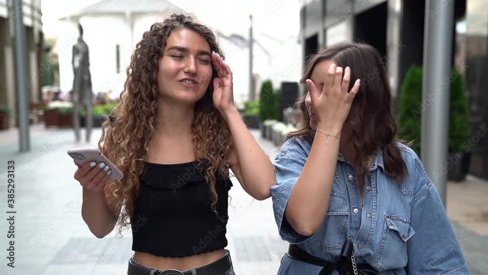girlfriends are walking along a city street and chatting. one curly-haired woman holds a phone and gestures, while the other listens girlfriends are walking along a city street and chatting. one curly-haired woman holds a phone and gestures, while the other listens