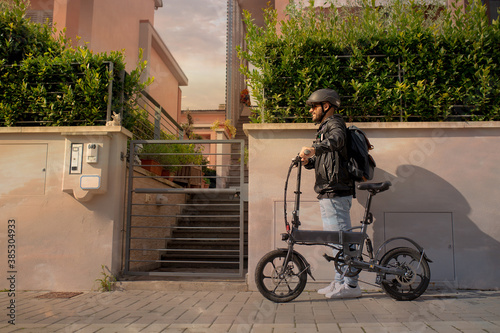 Smiling man with helmet and his electric bicycle at sunset. Commuter and new transportation concept. Green energy. Caucasian man drives his foldable e bike to go work or in leisure