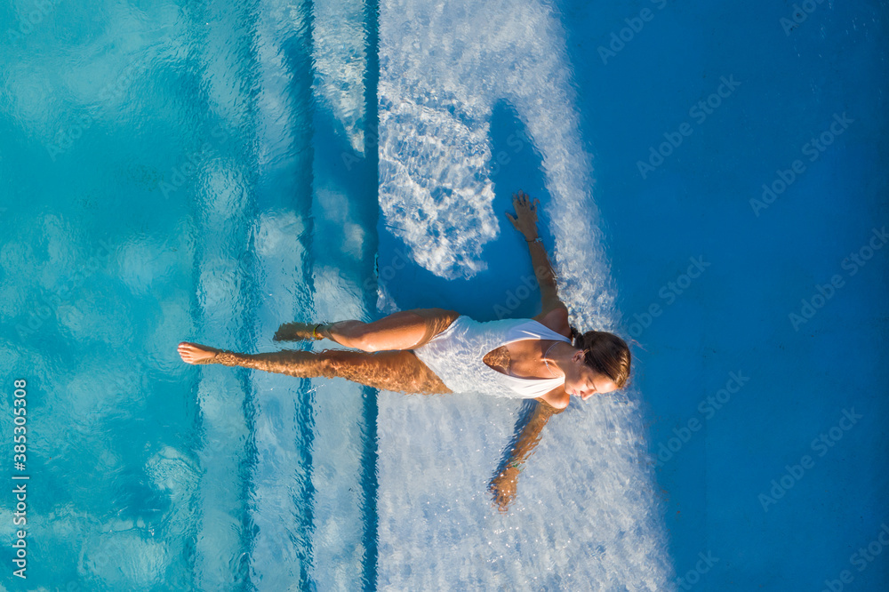 © AmazingAerialAgency - Aerial view of woman laying on the stairs of a swimming pool in a white swimsuit.