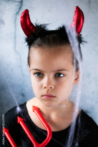 Portrait of little girl in Halloween costume of little devil with trident against the grey wall