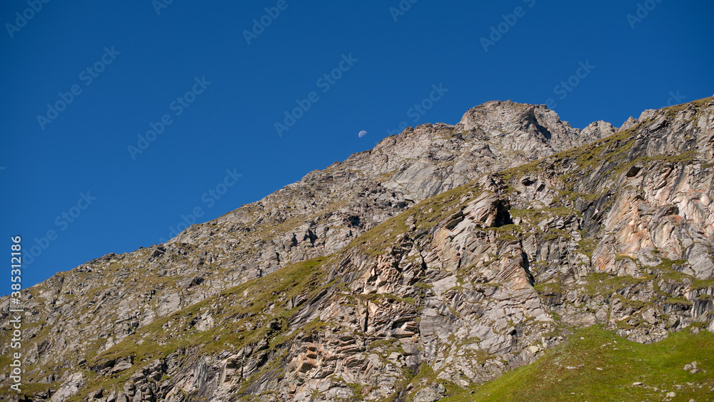 Mond am blauen Himmel über einer Felswand in den Hohen Tauern, Österreich