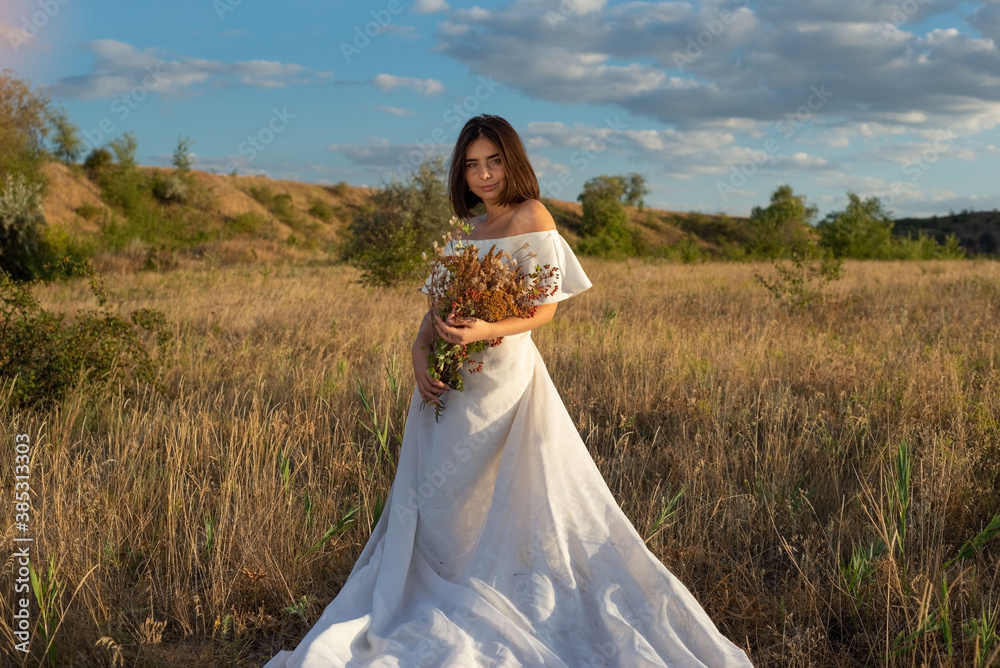 beautiful girl with flowers in the field. In a white wedding dress, standing in a field against the backdrop of a cloudy blue sky. Wedding outfit. A bouquet of wild flowers. beautiful girl with flowers in the field. In a white wedding dress, standing in a field against the backdrop of a cloudy blue sky. Wedding outfit. A bouquet of wild flowers.