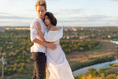 portrait of a young couple, a girl hugs a guy. In white clothes, in the field. Against the backdrop of the setting sun, cloudy blue sky. Warm tone photo. Love of a twenty two year old couple.