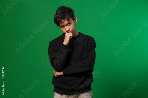 young asian man frowned on face looking at camera and hand on chin when make decisions. isolated on copy space green background for advertisement and design concept