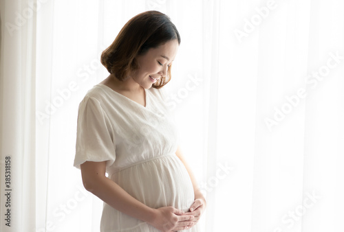 Asian pregnant woman wearing white dress and standing beside window
