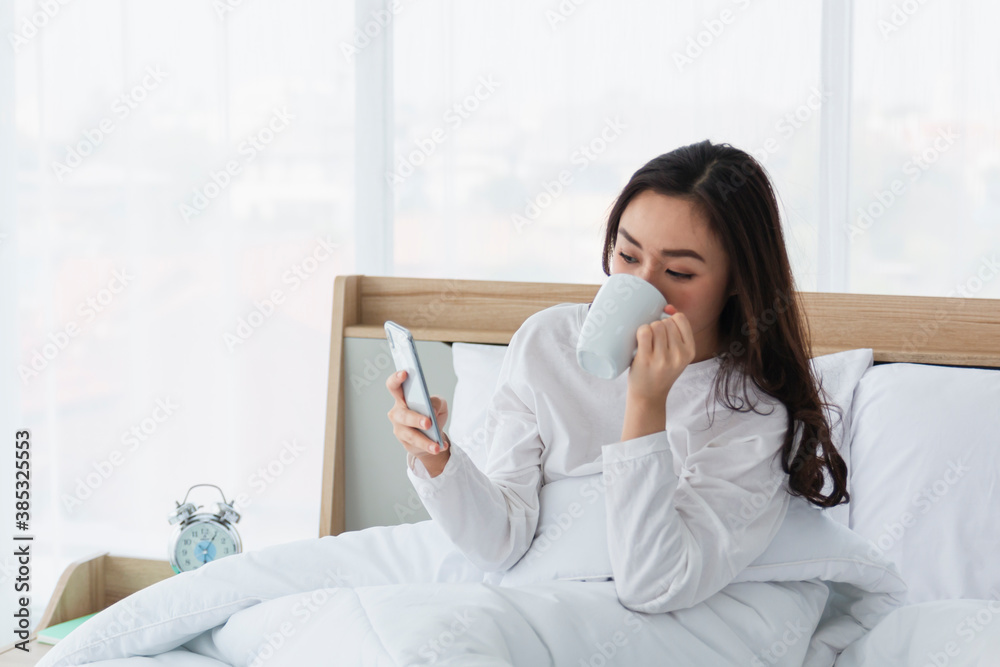 Young woman in white relaxing and drinking cup of hot coffee or tea using smartphone on bed in the bedroom. Happy girl using a mobile phone on bed. Beautiful woman using smartphone in the morning. Young woman in white relaxing and drinking cup of hot coffee or tea using smartphone on bed in the bedroom. Happy girl using a mobile phone on bed. Beautiful woman using smartphone in the morning.