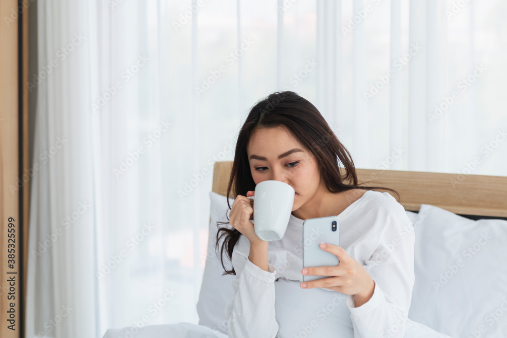 Young woman in white relaxing and drinking cup of hot coffee or tea using smartphone on bed in the bedroom. Happy girl using a mobile phone on bed. Beautiful woman using smartphone in the morning. Young woman in white relaxing and drinking cup of hot coffee or tea using smartphone on bed in the bedroom. Happy girl using a mobile phone on bed. Beautiful woman using smartphone in the morning.