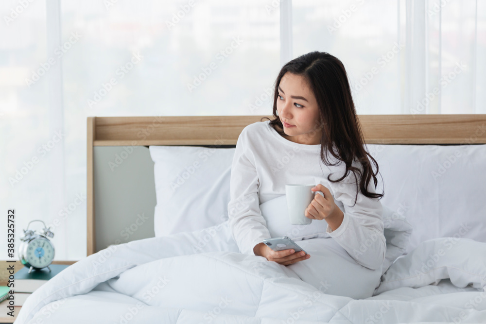 Young woman in white relaxing and drinking cup of hot coffee or tea using smartphone on bed in the bedroom. Happy girl using a mobile phone on bed. Beautiful woman using smartphone in the morning. Young woman in white relaxing and drinking cup of hot coffee or tea using smartphone on bed in the bedroom. Happy girl using a mobile phone on bed. Beautiful woman using smartphone in the morning.
