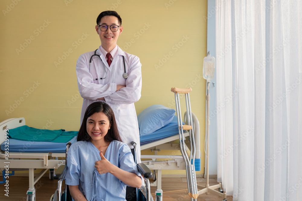 Fototapeta premium Healthcare and Treatment Concept. Young Asian female patient smiley face lifts hand up fighting with illness on wheelchair. Doctor and Patient showing fighting spirits over illness in hospital room.
