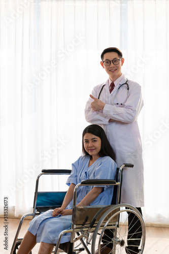 Healthcare and Treatment Concept. Young Asian female patient smiley face lifts hand up fighting with illness on wheelchair. Doctor and Patient showing fighting spirits over illness in hospital room.