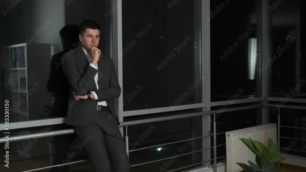 Pensive young man 30 year old businessman in suit standing in his office near a panoramic window overlooking the night city Pensive young man 30 year old businessman in suit standing in his office near a panoramic window overlooking the night city