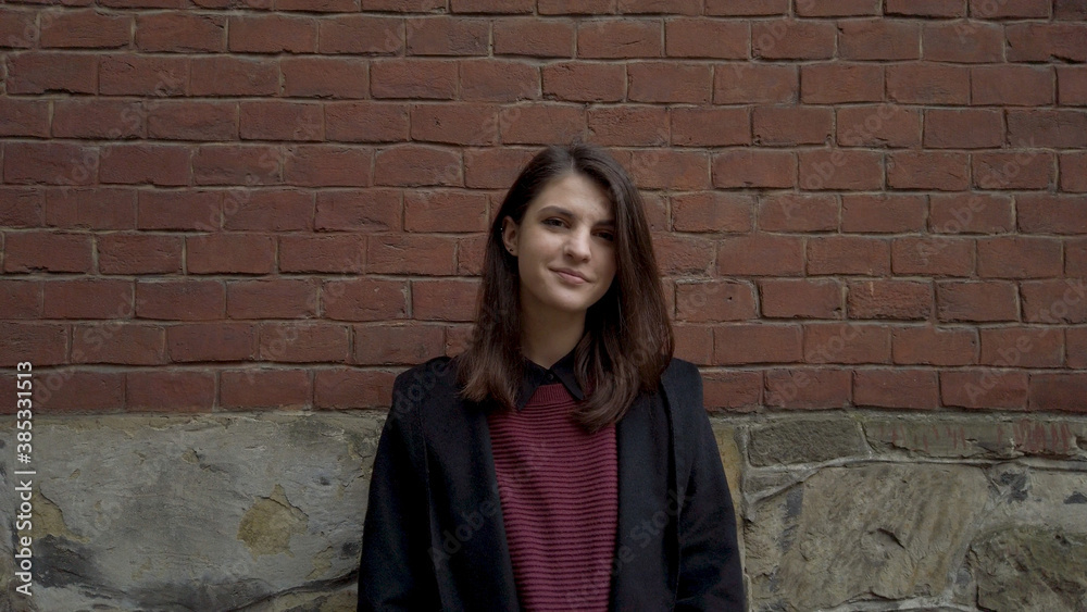 Portrait of cute young shy woman standing against red brick wall looking around and smiling for camera Portrait of cute young shy woman standing against red brick wall looking around and smiling for camera