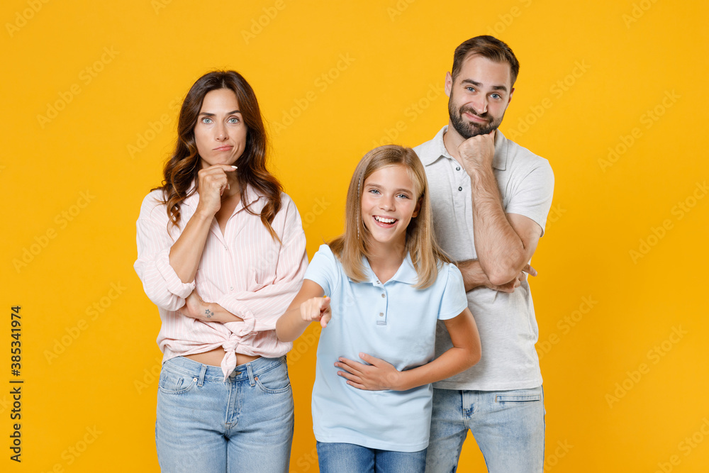 Fototapeta premium Confused young parents mom dad with laughing child kid daughter teen girl in t-shirts put hand prop up on chin pointing index finger on camera isolated on yellow background studio. Family day concept.