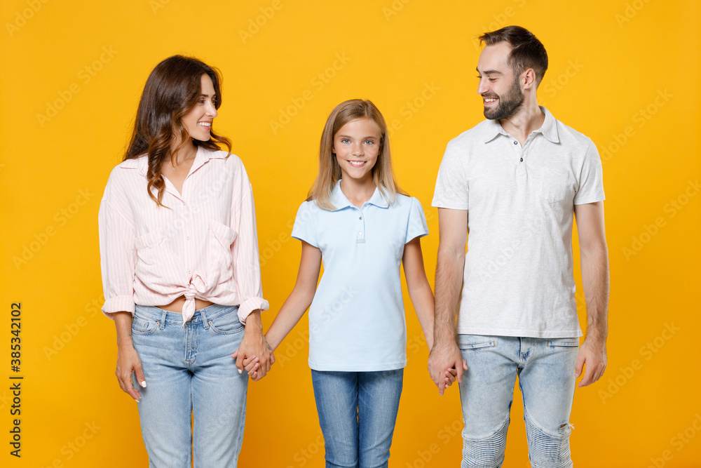 Smiling young parents mom dad child kid daughter teen girl in basic t-shirts holding hands looking at each other isolated on yellow background studio portrait. Family day parenthood childhood concept. Smiling young parents mom dad child kid daughter teen girl in basic t-shirts holding hands looking at each other isolated on yellow background studio portrait. Family day parenthood childhood concept.