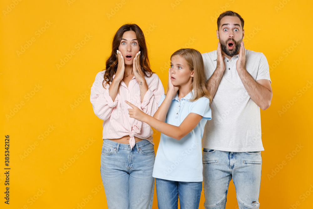 Shocked young parents mom dad with child kid daughter teen girl in basic t-shirts pointing index finger aside put hands on cheeks isolated on yellow background studio portrait. Family day concept. Shocked young parents mom dad with child kid daughter teen girl in basic t-shirts pointing index finger aside put hands on cheeks isolated on yellow background studio portrait. Family day concept.