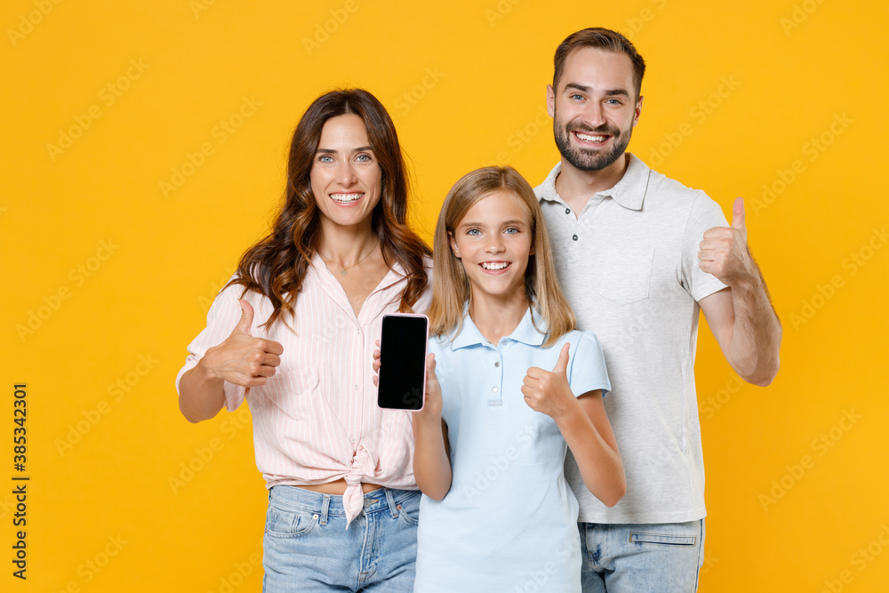 Obraz premium Smiling funny young parents mom dad with child kid daughter teen girl in basic t-shirts hold mobile phone with blank empty screen showing thumbs up isolated on yellow background. Family day concept.