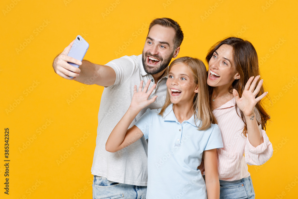 Funny young parents mom dad with child kid daughter teen girl in basic t-shirts doing selfie shot on mobile phone waving greeting with hands isolated on yellow background studio. Family day concept. Funny young parents mom dad with child kid daughter teen girl in basic t-shirts doing selfie shot on mobile phone waving greeting with hands isolated on yellow background studio. Family day concept.