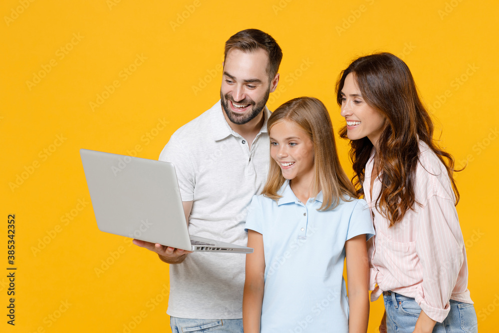 Smiling funny young parents mom dad with child kid daughter teen girl in basic t-shirts using laptop pc computer isolated on yellow background studio portrait. Family day parenthood childhood concept. Smiling funny young parents mom dad with child kid daughter teen girl in basic t-shirts using laptop pc computer isolated on yellow background studio portrait. Family day parenthood childhood concept.