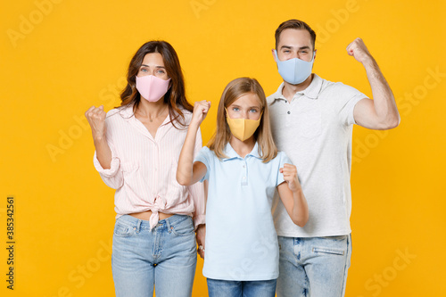 Happy young parents mom dad with child kid daughter teen girl in t-shirts face mask to safe from coronavirus virus covid-19 doing winner gesture isolated on yellow background. Family day concept.