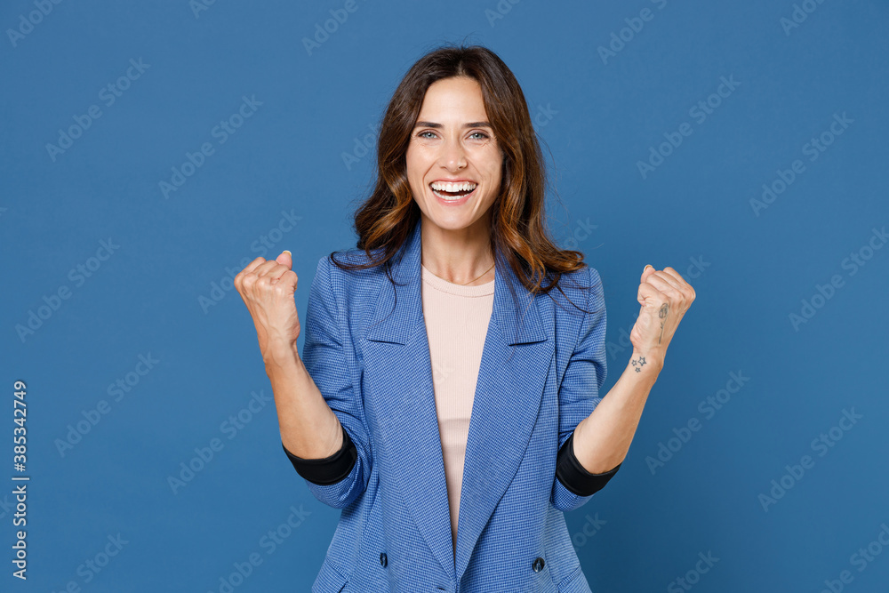 Happy joyful funny attractive young brunette woman 20s wearing basic jacket standing clenching fist doing winner gesture looking camera isolated on bright blue colour wall background studio portrait. Happy joyful funny attractive young brunette woman 20s wearing basic jacket standing clenching fist doing winner gesture looking camera isolated on bright blue colour wall background studio portrait.