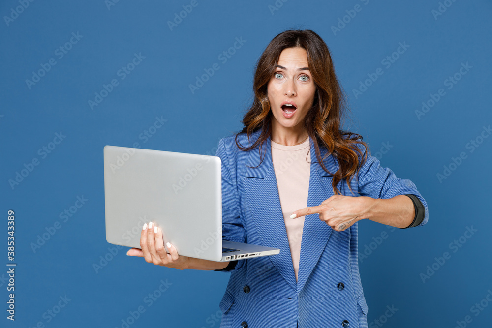 Shocked amazed young brunette woman 20s wearing basic jacket standing pointing index finger working on laptop pc computer looking camera isolated on bright blue colour wall background studio portrait. Shocked amazed young brunette woman 20s wearing basic jacket standing pointing index finger working on laptop pc computer looking camera isolated on bright blue colour wall background studio portrait.