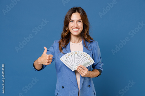 Obraz na plátně Smiling funny young brunette woman 20s wearing basic jacket standing holding in hand fan of cash money in dollar banknotes showing thumb up isolated on bright blue colour background studio portrait