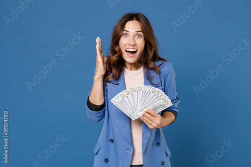 Obraz na plátně Surprised amazed young brunette woman 20s wearing basic jacket standing holding in hand fan of cash money in dollar banknotes spreading hands isolated on bright blue colour background studio portrait