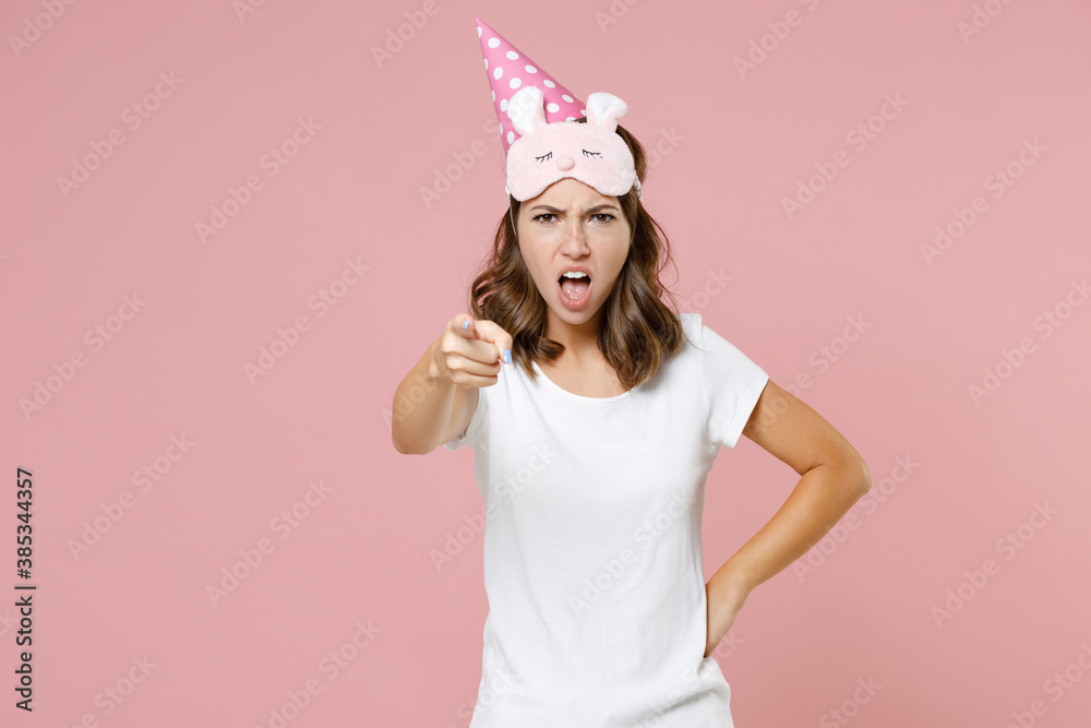 Angry young woman in pajamas home wear sleep mask birthday hat point index finger on camera swearing rest at home isolated on pastel pink background studio portrait. Relax good mood lifestyle concept. Angry young woman in pajamas home wear sleep mask birthday hat point index finger on camera swearing rest at home isolated on pastel pink background studio portrait. Relax good mood lifestyle concept.
