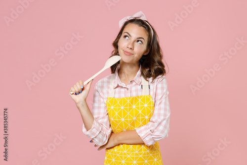 Pensive young woman housewife 20s wearing yellow apron checkered shirt hold spoon soup ladle dipper looking up doing housework isolated on pastel pink background studio portrait. Housekeeping concept.