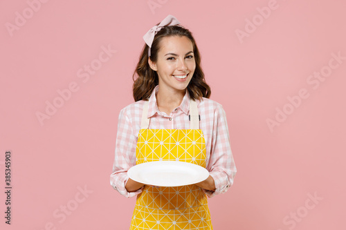 Beautiful smiling young brunette woman housewife 20s in yellow apron checkered shirt hold empty plate while doing housework isolated on pastel pink background studio portrait. Housekeeping concept. photo