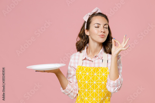Fotografie Cute young woman housewife in yellow apron hold empty plate making okay taste delight sign keeping eyes closed doing housework isolated on pastel pink background studio portrait
