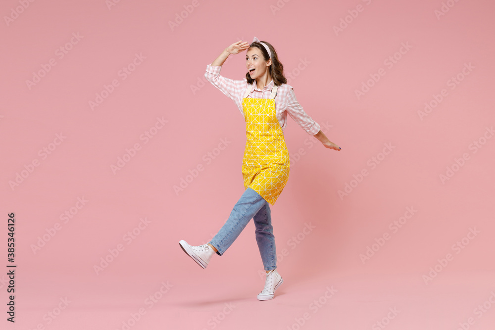 Full length portrait excited young woman housewife in apron holding hand at forehead looking far away distance doing housework isolated on pastel pink colour background studio. Housekeeping concept.