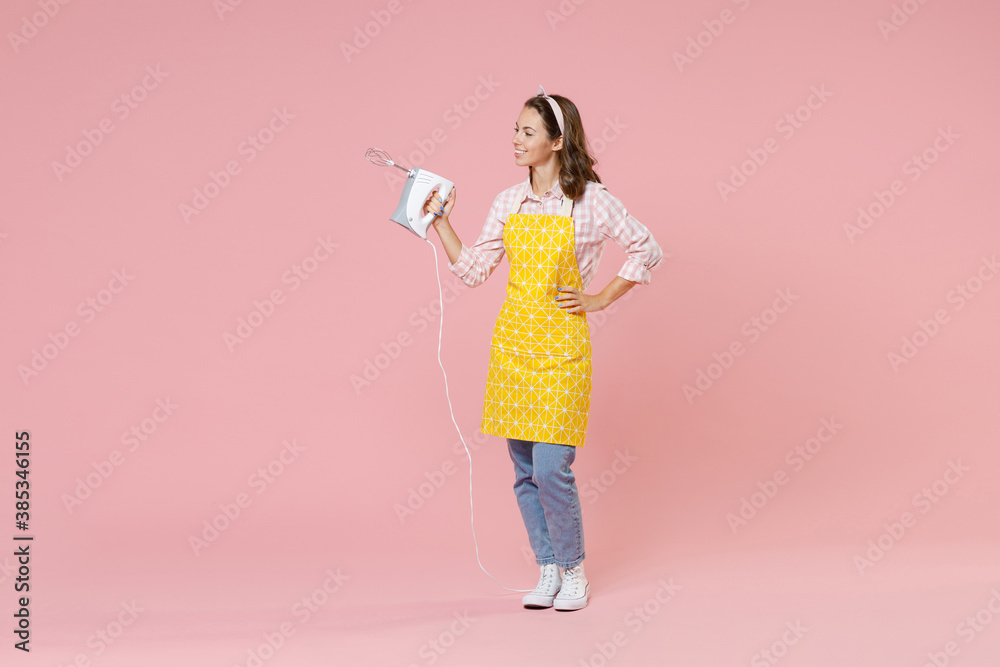 Full length portrait of smiling pretty young brunette woman housewife 20s wearing yellow apron hold mixer while doing housework isolated on pastel pink colour background studio. Housekeeping concept. Full length portrait of smiling pretty young brunette woman housewife 20s wearing yellow apron hold mixer while doing housework isolated on pastel pink colour background studio. Housekeeping concept.