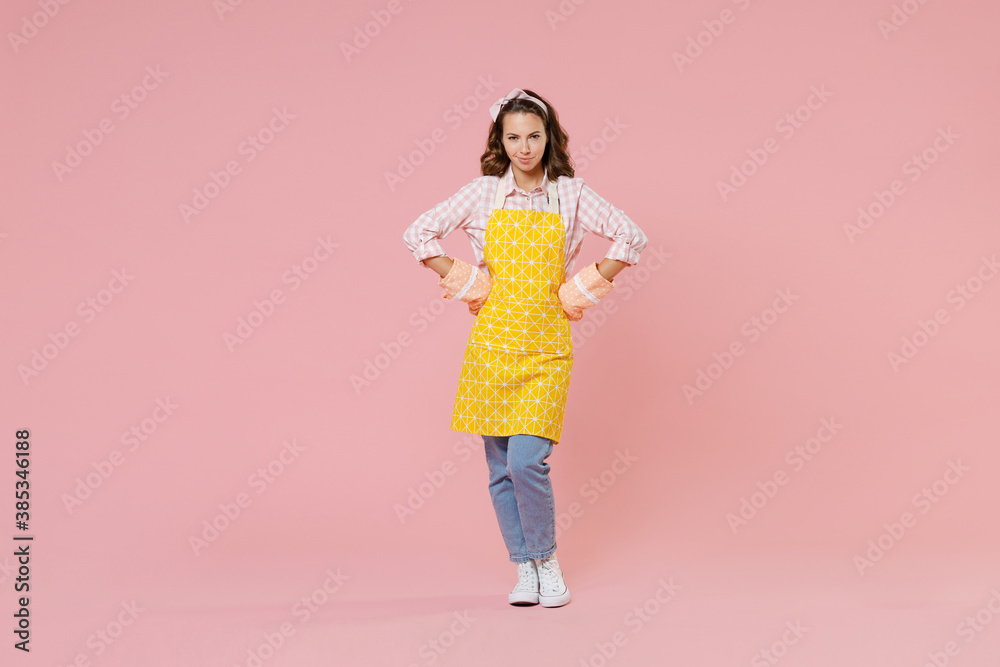 Full length portrait pretty young woman housewife in apron gloves potholders standing with arms akimbo on waist doing housework isolated on pastel pink colour background studio. Housekeeping concept. Full length portrait pretty young woman housewife in apron gloves potholders standing with arms akimbo on waist doing housework isolated on pastel pink colour background studio. Housekeeping concept.