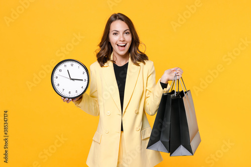 Excited young brunette woman wearing basic light suit jacket hold package bags with purchases after shopping clock looking camera isolated on yellow background, studio portrait. Black friday sale.