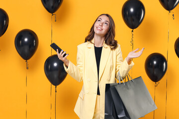 Smiling pensive young woman in suit jacket hold package bags with purchases after shopping using mobile phone looking up on yellow background with air balloons studio portrait. Black friday sale.