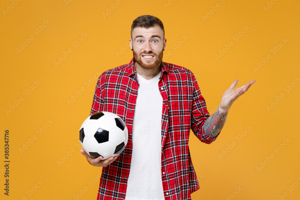Confused puzzled young man football fan in basic red shirt cheer up support favorite team with soccer ball spreading hands isolated on yellow background studio. People sport leisure lifestyle concept. Confused puzzled young man football fan in basic red shirt cheer up support favorite team with soccer ball spreading hands isolated on yellow background studio. People sport leisure lifestyle concept.